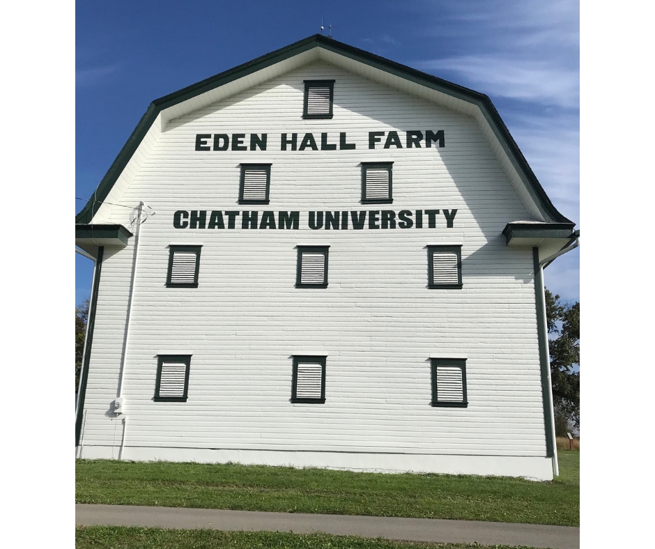 A white barn with a blue sky behind it. It sits on a plot of green grass. The barn has the words Eden Hall Farm Chatham University placed on it in block letters