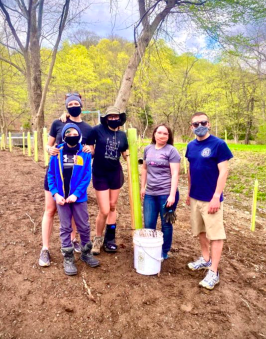 Group photo of Claire with other volunteers, all persons masked and standing with tools used to plant trees.  Saplings that have already been planted are in the background.