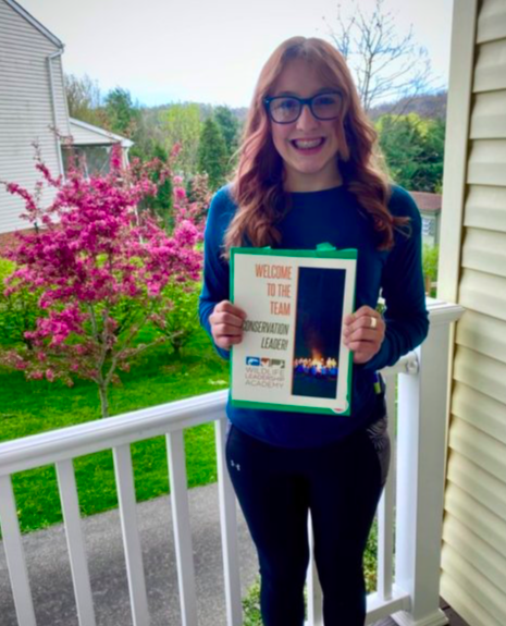 Claire stands smiling on her porch, holding her acceptance certificate to the Wildlife Leadership Academy, which reads "Welcome to the team!"