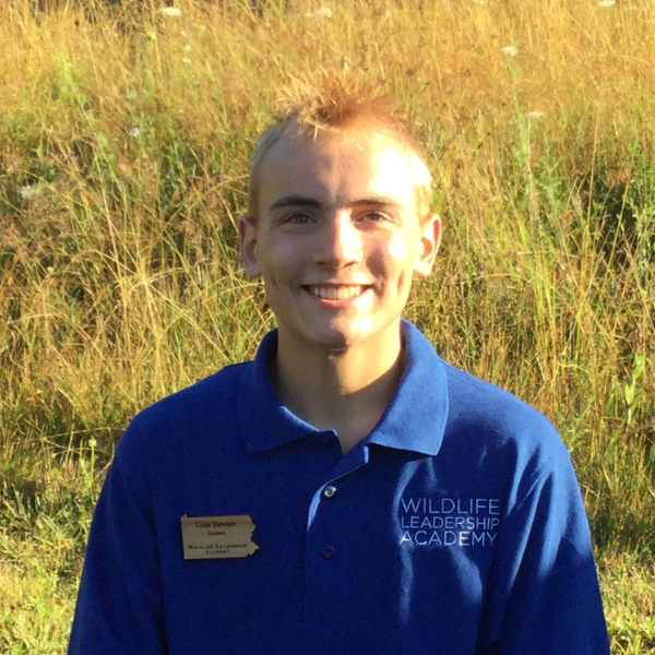 Image of a young man, Cole, wearing a royal blue polo shirt embroidered with the words "Wildlife Leadership Academy" and a Pennsylvania state shaped brown wooden nametag.