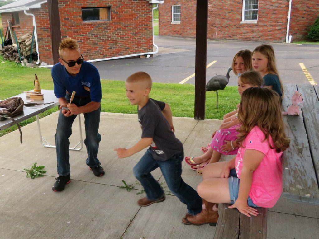 Image of Cole using a slate turkey call to imitate a turkey while a young child runs nearby, pretending to be a turkey.  Other young children sit nearby, observing Cole and the young child.